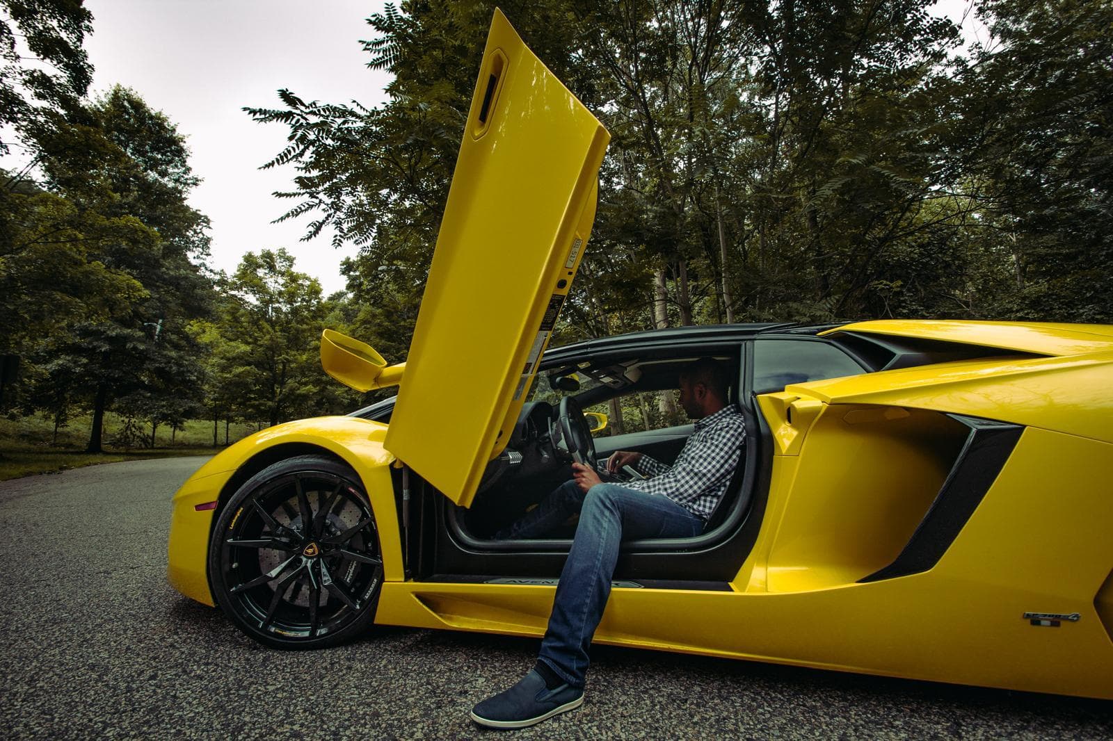 Driver seated in a yellow Lamborghini Aventador Roadster with its scissor door raised, side view